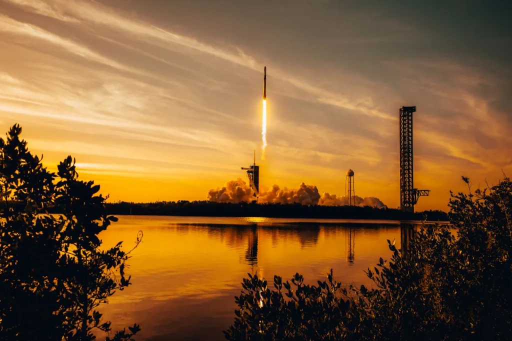 A rocket lifts off with the view from across a body of water at dawn